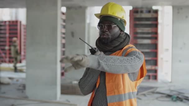ハードハットとセーフティーベストを身に着けている黒人男性建設現場のスーパーバイザーのミディアムショットは ウォルキーのトーキーに作業員に指示を与えながら 手のジェスチャーを作ります — ストック動画