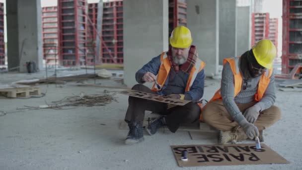 未完成の建設現場のコンクリートフロアに座り 一緒にストライキする前に段ボールバナーに抗議スローガンを描いた2人の多民族建設労働者 — ストック動画