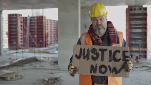 ダンボールジャスティス今ストライキバナーと建設現場に立っている深刻な中年白人男性建設労働者の中程度のスローモの肖像 — ストック動画