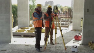 Açıyı ölçmek ve tartışmak için bütün istasyon boyunca sabit şapkalı ve yansıtıcı yelekli iki farklı mühendisin beton şantiyede birlikte çalışması uzun bir çekim.