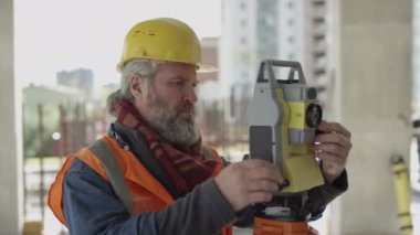 Profesyonel orta yaşlı, sarı başlıklı beyaz mühendis, tamamlanmamış inşaat sahasında çalışırken tüm istasyonu kullanıyor.
