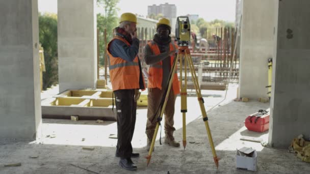 两名戴着硬礼帽 身穿反光安全背心 站在总站旁进行角度测量和讨论 在混凝土建筑工地一起工作的工程师的长篇全景照片 — 图库视频影像