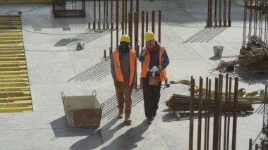 Yukarıdan çekilen iki farklı işçinin portakal rengi yansıtıcı yelekleri ve kalın şapkaları ile bitmemiş inşaat alanının beton çatısında sohbet ederken görüntüsü.