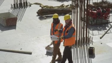 İki çok ırklı inşaat mühendisinin güneşli bir günde beton binanın çatısının planlarını tartışırken yüksek açılı görüntüsü.