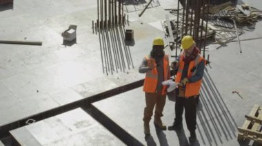 İki profesyonel inşaat mühendisinin, inşaat halindeki çok katlı beton emlak binasının çatısında dururken inşaat planlarını tartışırken yukarıdan çekilen görüntüleri.