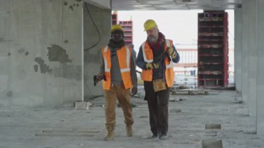 Beton inşaat sahasında yürürken ve sohbet ederken iki farklı etnik kökenli işçi el aletleri tutuyor.