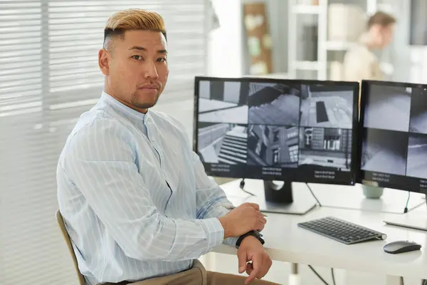 Side view portrait of young Asian man looking at camera with surveillance feeds in background, security center office, copy space
