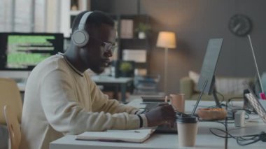Yan görüş, genç Afrikalı Amerikalı erkek programcının kablosuz kulaklık ve gözlük takıp minimalist ofiste dizüstü bilgisayarla çalışırken görüntüsü.