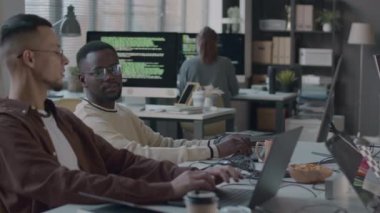 Modern ofisteki bir masada oturan, dizüstü bilgisayarda yazan ve arka planda çalışan iki farklı erkek programcının belini kaldırın.