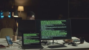 Karanlık ofisteki masada bilgisayar ve dizüstü bilgisayar üzerindeki html kodlarının satırları. İki erkek programcı işyerlerinde oturuyor ve yeni yazılımlar geliştiriyor.