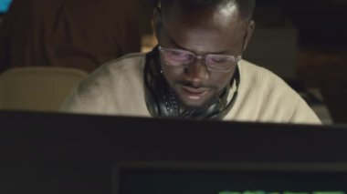 Karanlık ofiste bilgisayarın önünde oturan konsantre genç siyahi erkek yazılım mühendisinin orta ölçekli yakın çekimi ve yeni bir program geliştirilmesi.