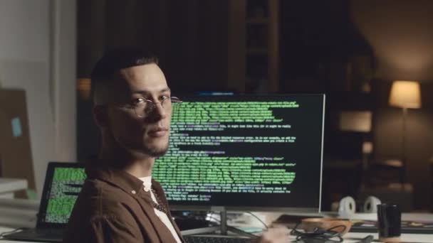 若いBiracialソフトウェアエンジニアのミディアム肖像画 鼻に眼鏡を調整し 暗いオフィスの黒い画面でHtmlコードでデスクトップコンピュータに座っている間カメラを見ています — ストック動画