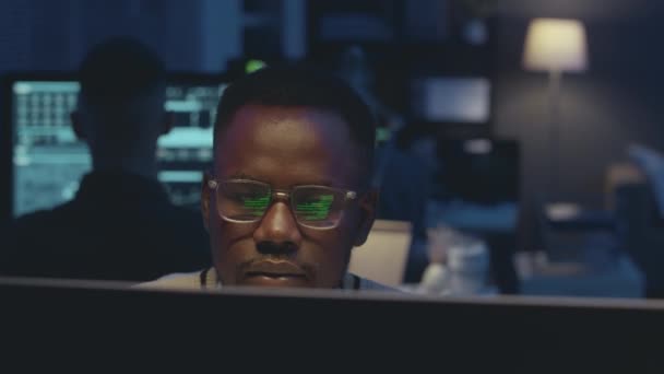 若い黒人男性のサイバーセキュリティワーカーは夜に暗いオフィスでコンピュータで作業し 黒い背景の緑のプログラムコードを眼鏡に反映しています — ストック動画