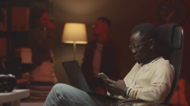Genç siyahi erkek yazılım geliştiricisinin, ofis koltuğunda dizüstü bilgisayarla otururken arka planda çeşitli meslektaşlarıyla sohbet ederken orta boy görüntüsü.