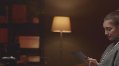 Beyaz kadın bilişim koordinatörü ve dijital tabletinin belden yukarı portresi. Kameraya gülümsüyor. Gece ofisindeki ofis sandalyesinde kırmızı neon ışıkla oturuyor.