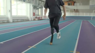 Kapalı stadyumdaki pistte bıçakla koşan kesik bacaklı beyaz erkek para-sporcunun eğik çekimini izliyorum.