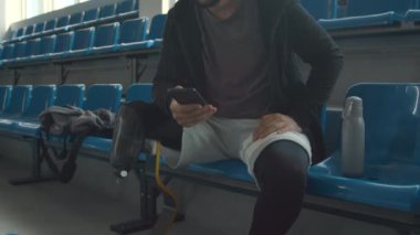 Beyaz tenli sporcunun spor salondaki tribün koltuğunda antreman yaptıktan sonra dinlenen, su içen, akıllı telefon kullanan ve kablosuz kulaklıkla müzik dinleyen orta boy görüntüsü.