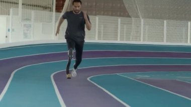 Kapalı stadyumda protez bacaklı beyaz erkek sporcu yavaş yavaş koşuyor.