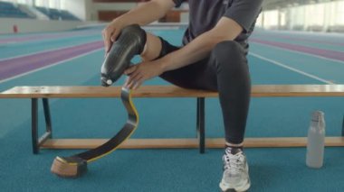 Ampüte edilmiş uzvundan protez koşu bıçağını çıkaran beyaz erkek sporcu kapalı stadyumdaki ahşap bankta otururken su içiyor ve antrenmandan sonra dinleniyor.