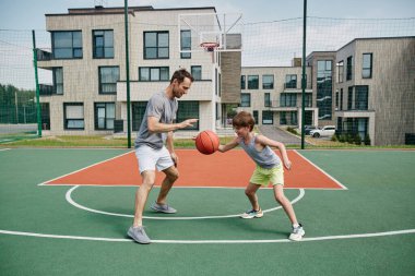 Baba ve oğlunun modern konut kompleksi tarafından açık havada basketbol oynarken geniş açılı çekim.