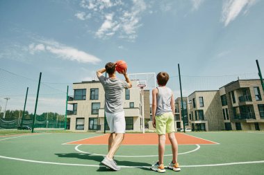 Baba ve oğlunun açık havada basketbol oynayıp potaya top attıkları geniş açılı çekim.