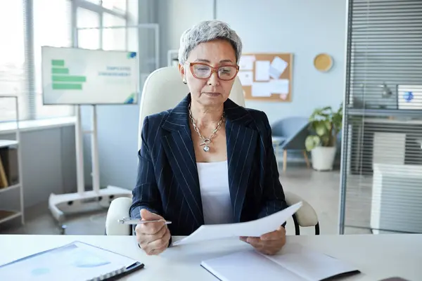 Portrait of senior woman as female boss sitting at workplace in office and reading document