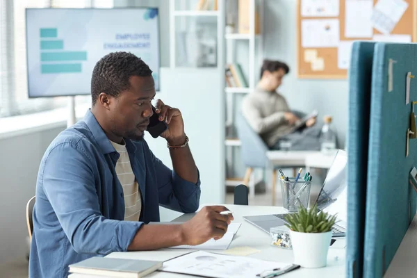 Side view portrait of black man speaking on phone while working at desk in office, copy space