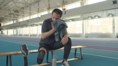 Sakallı, protez bacaklı beyaz bir adam antrenmandan sonra kapalı stadyumdaki ahşap bankta dinlenirken alnından terini havluyla siliyor.