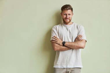 Kameraya gülümseyen sakallı genç adamın minimum portresi. Kolları pastel yeşil duvara çapraz durduğunda, boşluğu kopyala.