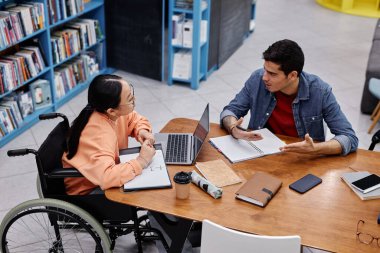 Engelli Asyalı genç kızın yüksek açılı portresi. Üniversite kütüphanesinde birlikte okuyorken arkadaşıyla konuşuyor.