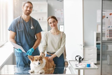 Corgi Dog 'un ve üniformalı erkek veterinerin mutlu genç sahibi şirin bir hayvanla sağlık masasının yanında dururken kameraya bakıyor.