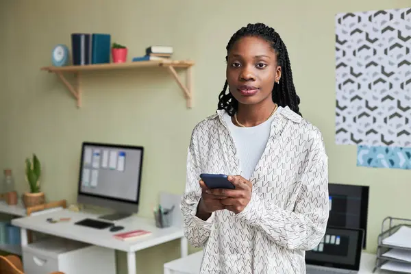 Waist up portrait of young black woman using smartphone and looking at camera while taking break in cozy office setting, copy space