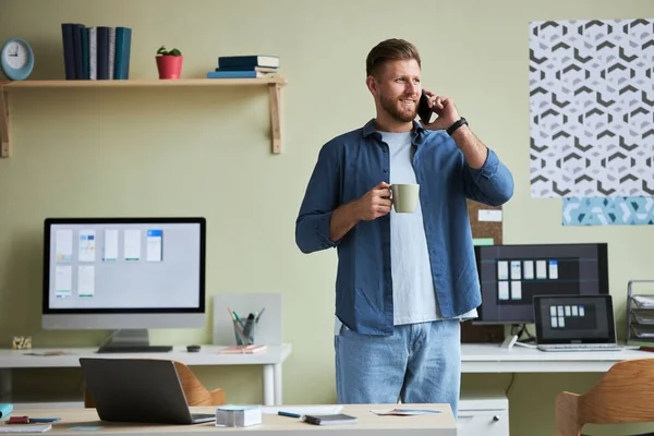 Portrait of smiling young man calling by phone and enjoying coffee while taking break from work in office, copy space