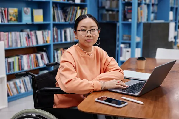 Portrait of Asian young woman with disability as college student looking at camera in colorful library setting with laptop, copy space