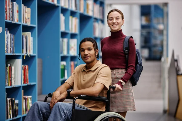 Portrait of smiling young woman assisting student with disability in library both looking at camera