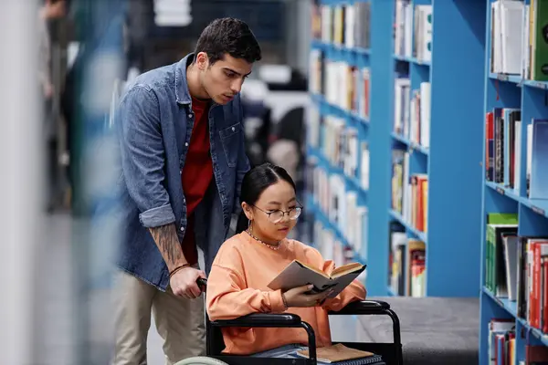 Portrait of Asian girl with disability in library choosing books with friend assisting, inclusivity concept