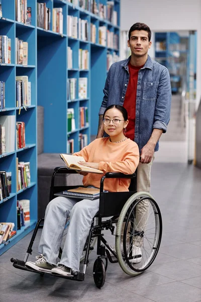 Full length portrait of smiling young man assisting female student with disability in library both looking at camera