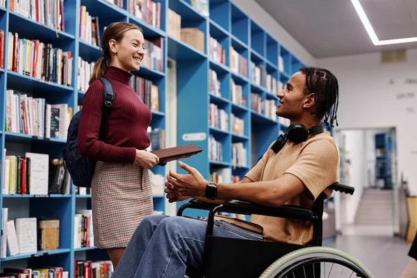 Side view portrait of black young student with disability talking to friend in college library and smiling