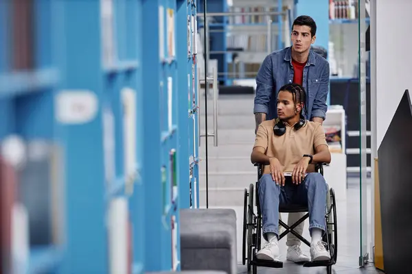 Graphic full length portrait of male student with disability using wheelchair in library with friend assisting, copy space