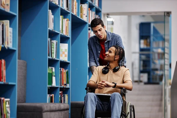 Portrait of smiling student with disability using wheelchair in library with friend assisting, copy space