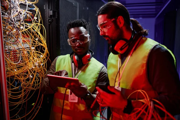 Close up of two network technicians using tablet while repairing server in neon light