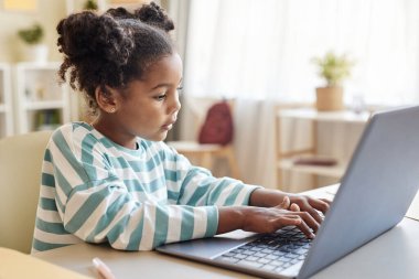 Evde internetten çalışırken dizüstü bilgisayar kullanan sevimli siyah kızın yan görünüm portresi