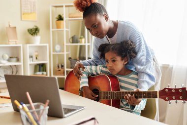 Annesiyle online müzik dersi izleyen ve evde gitar çalan siyah bir kızın portresi.