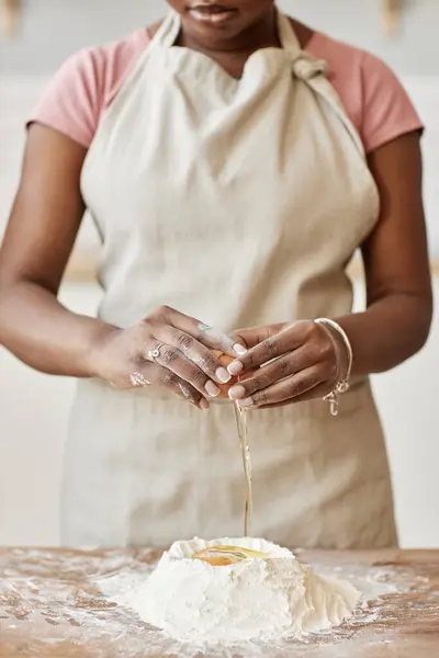 Minimal cropped portrait of black woman baking in kitchen and wearing apron while breaking eggs and mixing with flour making dough