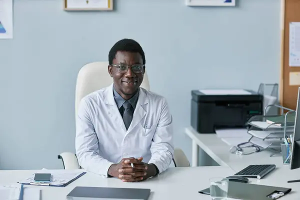 Minimal front view portrait of smiling black doctor looking at camera while sitting at desk in clinic office, copy space