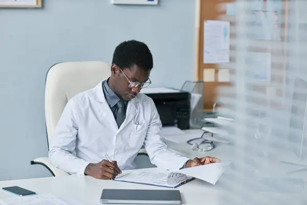 Portrait of young black doctor filling papers at desk in office while working in private clinic