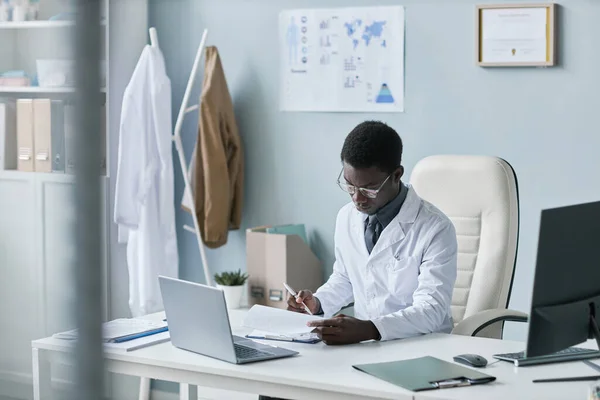 Portrait of young black doctor working with documents at desk in office and wearing lab coat