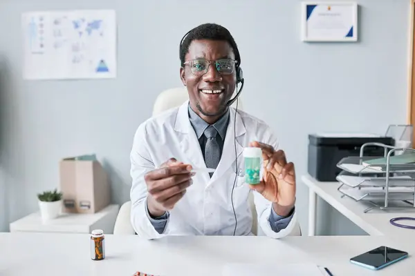 Portrait of black doctor holding bottle of pills and wearing headset at workplace in office