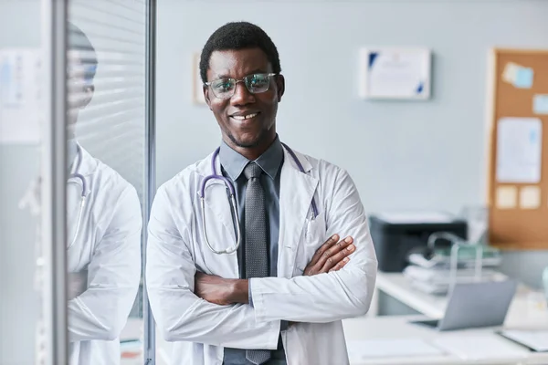 Waist up portrait of confident black doctor leaning on glass wall in office and smiling at camera, copy space