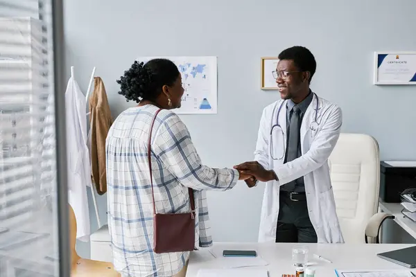 Portrait of smiling black doctor holding hands with female patient during friendly greeting for consultation in private clinic